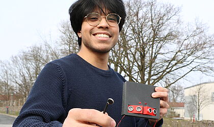 ein lächelnder junger Mann, Mustafa Rahmati, hält einen kleinen grauen Kasten mit vier lautsprecherähnlichen Sensoren und roten Kabeln